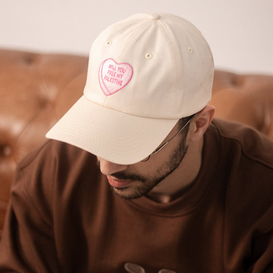 Man sat on sofa wearing a off white dad hat with a pink heart, similar to candy hearts, with the text 'will you free my Palestine'.