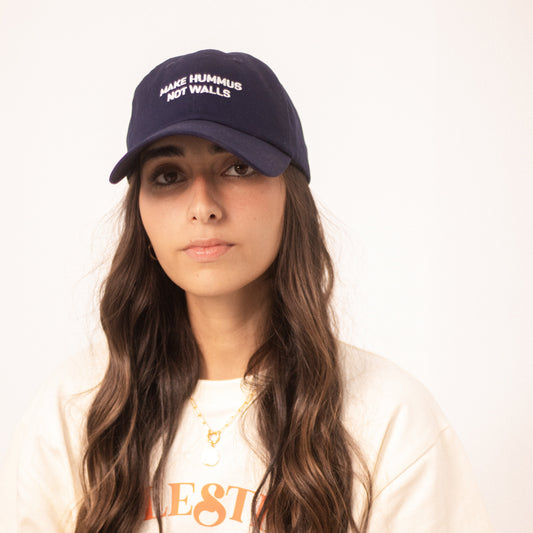 Image shows a model wearing a blue dad hat with with the text 'make hummus not walls' embroidered in white.