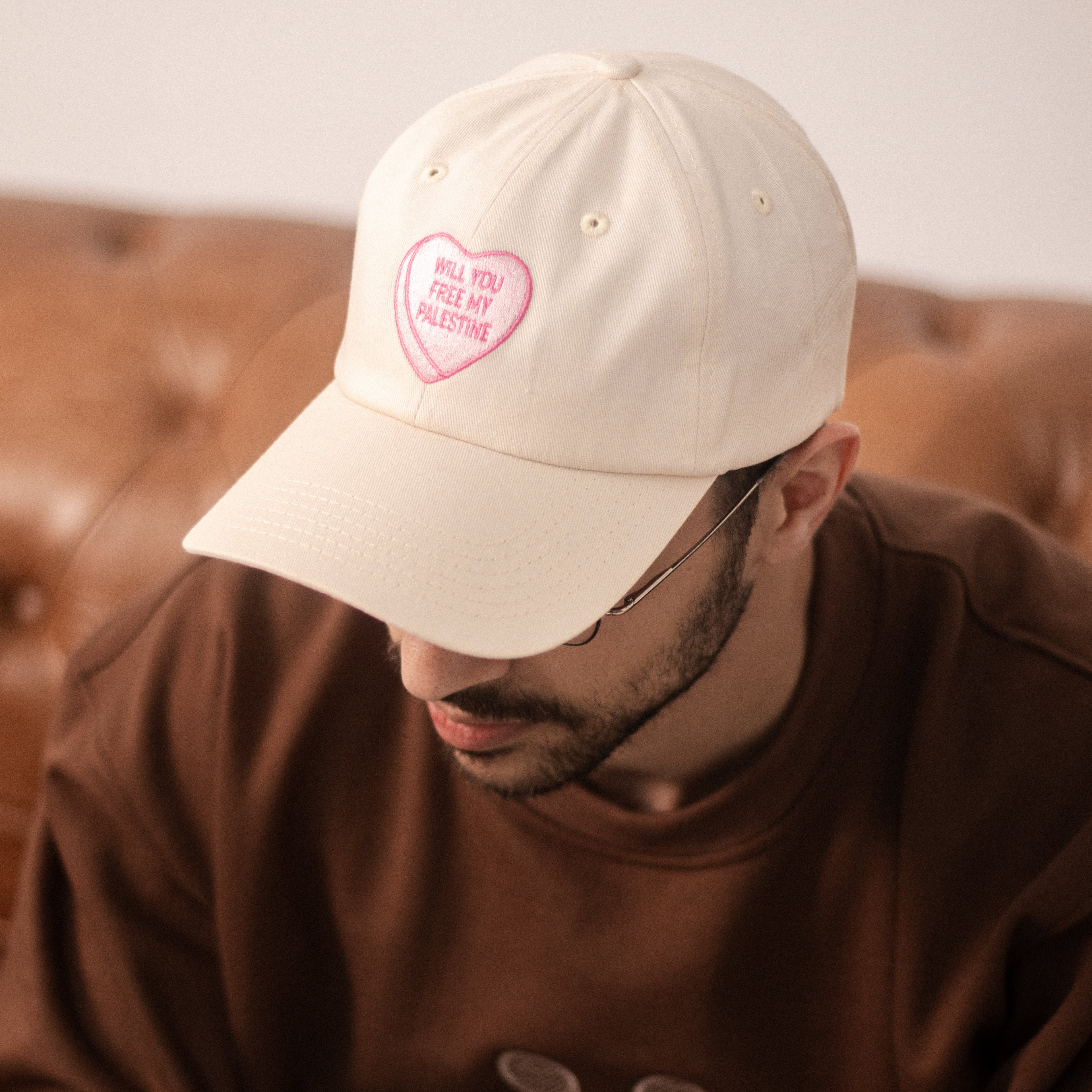Man sat on sofa wearing a off white dad hat with a pink heart, similar to candy hearts, with the text 'will you free my Palestine'. 
