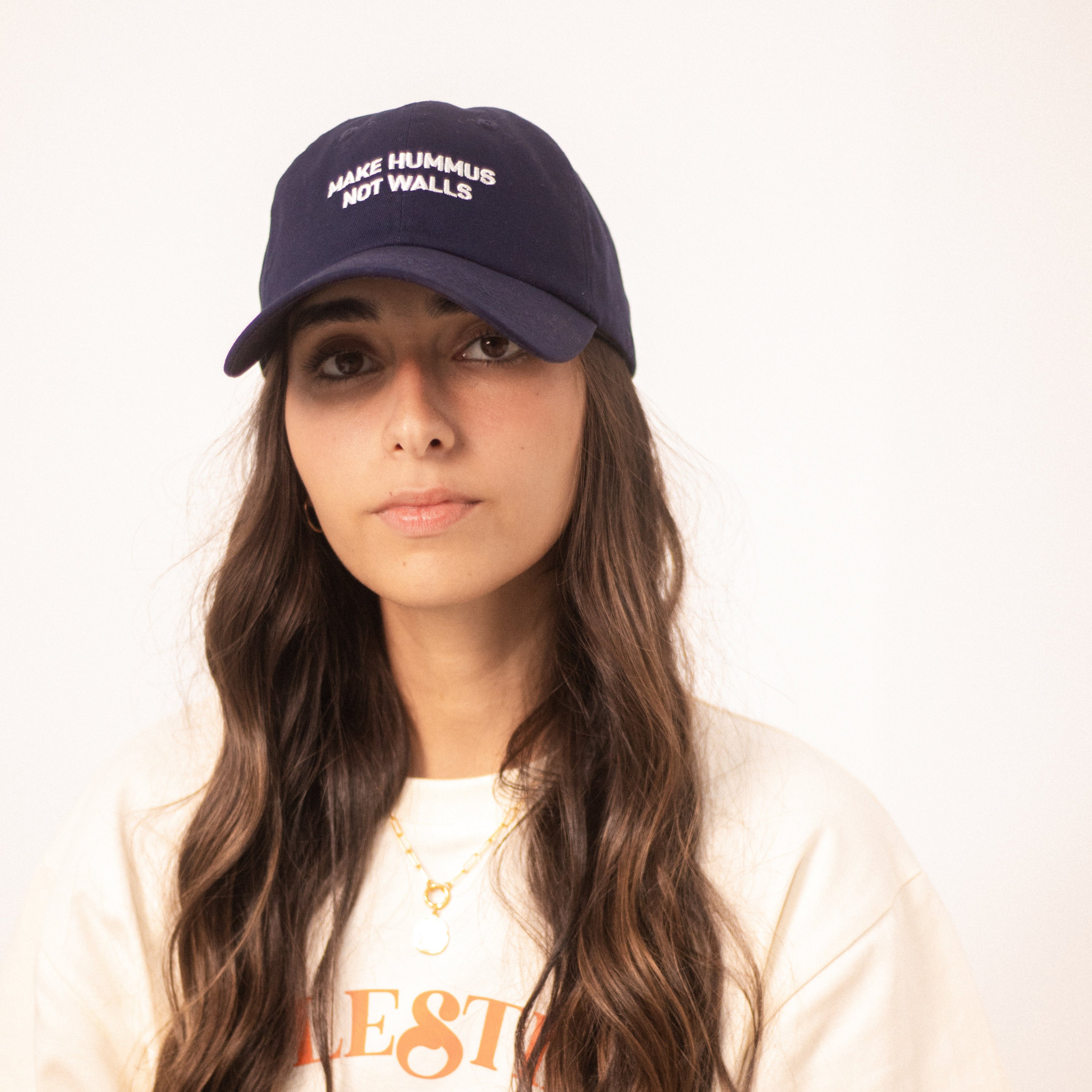 Image shows a model wearing a blue dad hat with with the text 'make hummus not walls' embroidered in white.