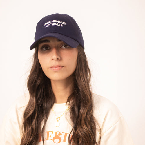 Image shows a model wearing a blue dad hat with with the text 'make hummus not walls' embroidered in white.